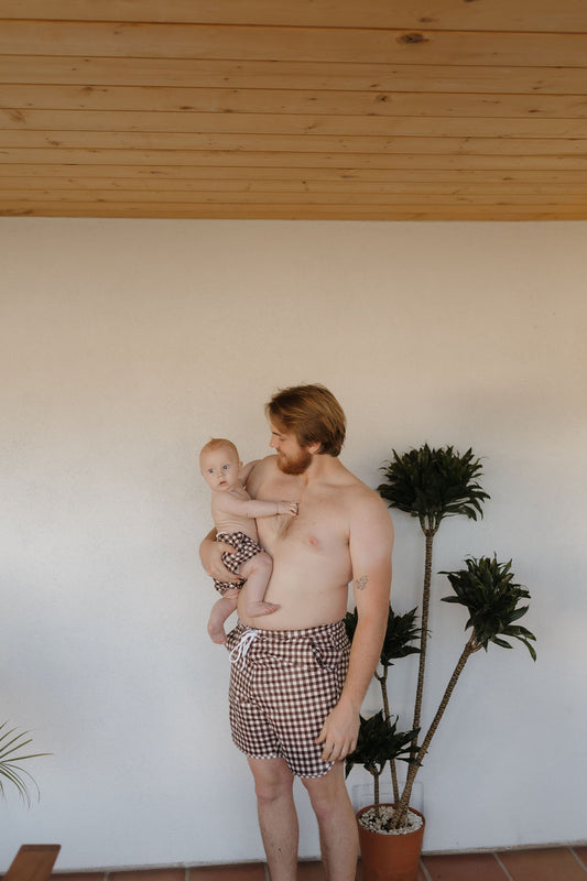 A shirtless man indoors wears Men's Boardshorts in Chocolate Gingham, matching his child's Forever French Baby outfit. They pose beside a potted plant under a wooden ceiling against light walls.