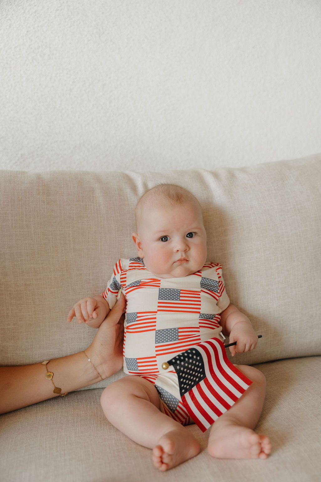 A baby in the Forever French Baby Bamboo Short Set | Americana sits on a light-colored couch, holding a small American flag while an adult’s hand gently supports them.