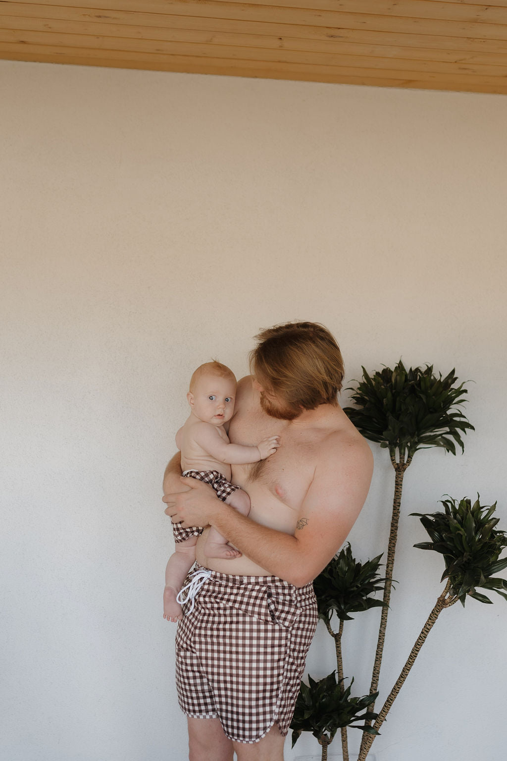 A man and baby, wearing matching Men's Boardshorts in Chocolate Gingham by forever french baby, stand near a minimalist backdrop with a plant. The man gazes affectionately at the baby, capturing the bright and sweet charm of Forever French Baby.