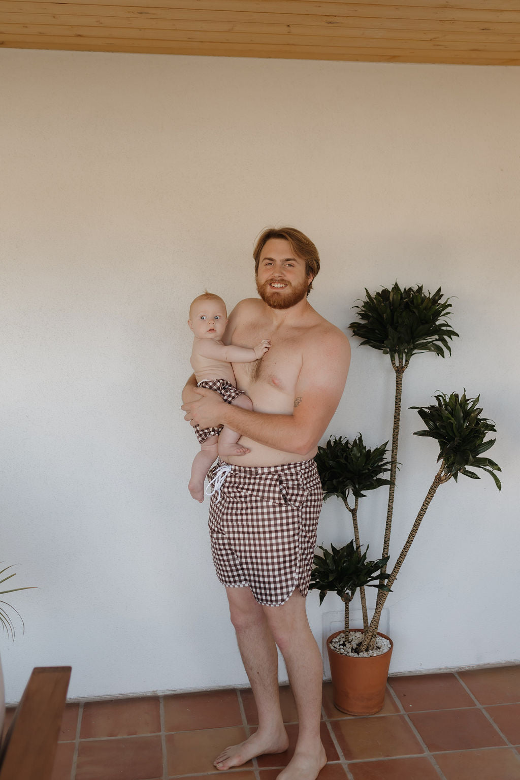 A shirtless man with a beard holds a baby, both wearing matching Men's Boardshorts in Chocolate Gingham by forever french baby. They stand on a tiled floor next to a potted plant, against a plain white wall, showcasing the timeless style of the collection.