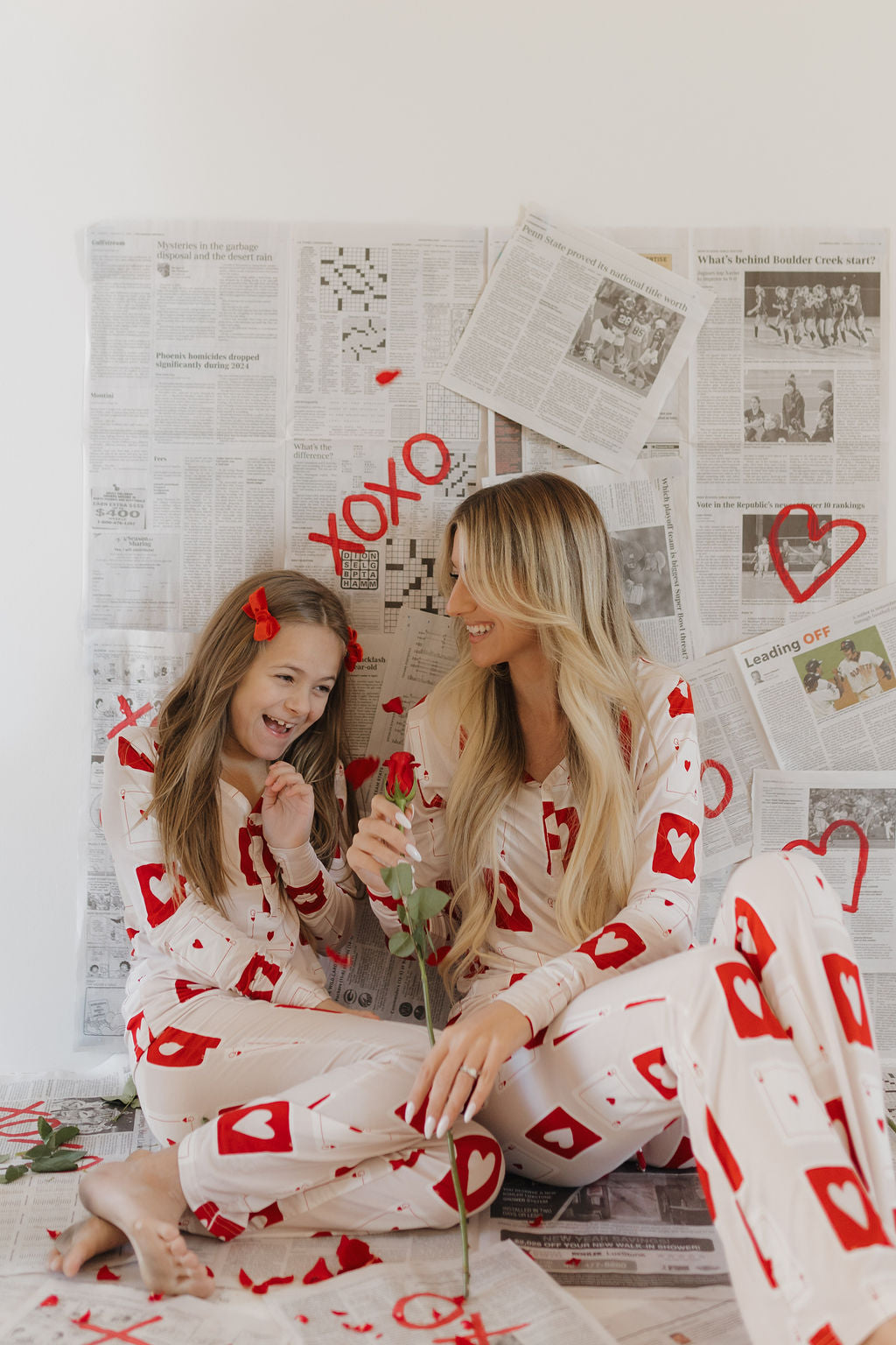 A woman and girl in matching "Youth Flare Bamboo Pajamas | Love Day" from forever french baby sit on the floor with newspaper pages around them. The woman holds a rose as the girl smiles, with "XOXO" on the wall, highlighting their cozy, hypo-allergenic outfit choice.