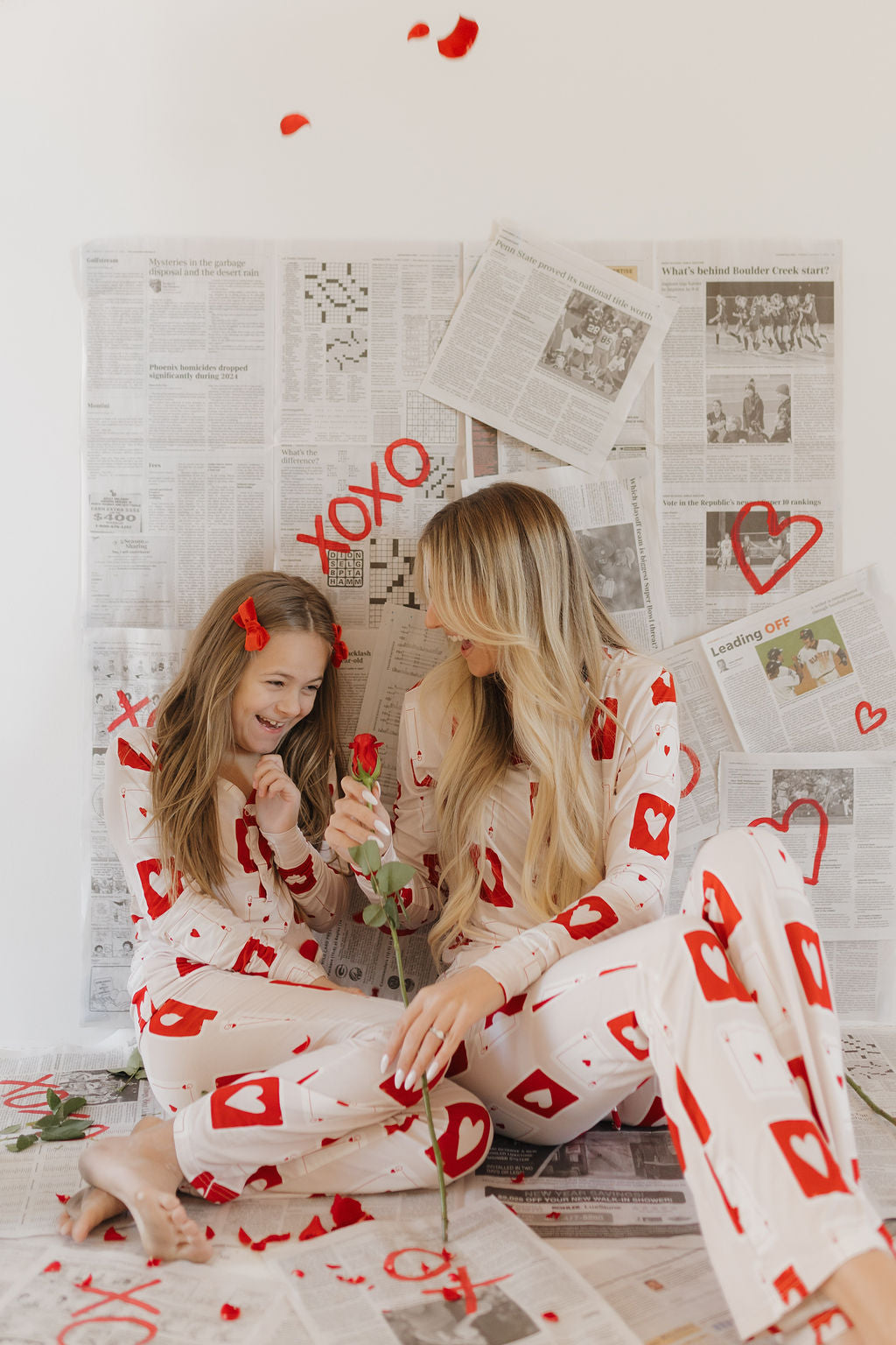 In forever french baby's Youth Flare Bamboo Pajamas | Love Day, two people sit on a newspaper-clad floor with red hearts and "XOXO," smiling amidst rose petals. The matching breathable bamboo fabric ensures comfort as they hold roses, adding to the romantic scene.