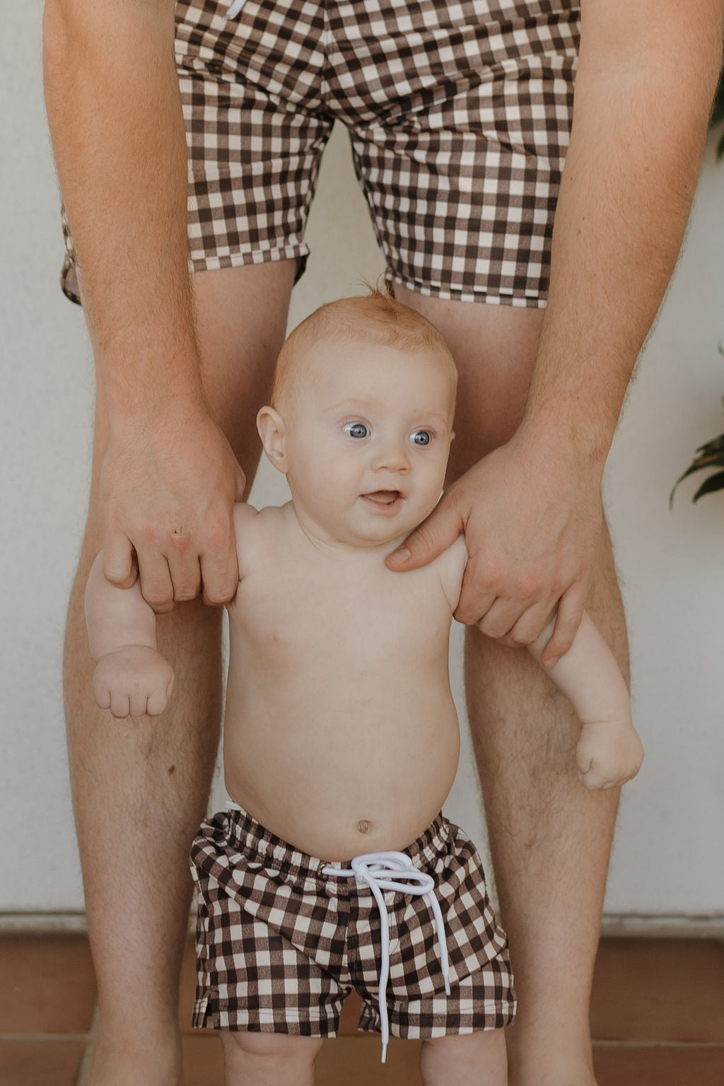 An adult lifts a baby from the Chocolate Gingham collection, both in matching brown and white checkered Men's Boardshorts by Forever French Baby. The baby's surprised expression adds charm to their coordinated look against a neutral background.