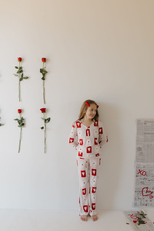 Against a white wall, a young girl stands barefoot in forever french baby's Youth Flare Bamboo Pajamas | Love Day, featuring red card suit symbols. Six red roses line the wall vertically, while newspapers with drawings and text are scattered on the floor nearby.