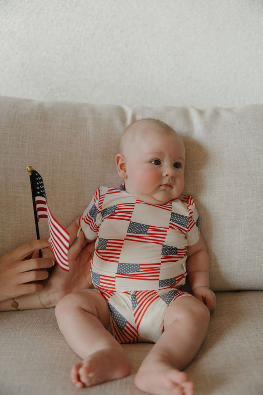 A baby sits on a beige couch wearing the forever french baby Bamboo Short Set | Americana, decorated with American flags. An adult hand beside the baby holds a small U.S. flag as the calm baby gazes to the side.