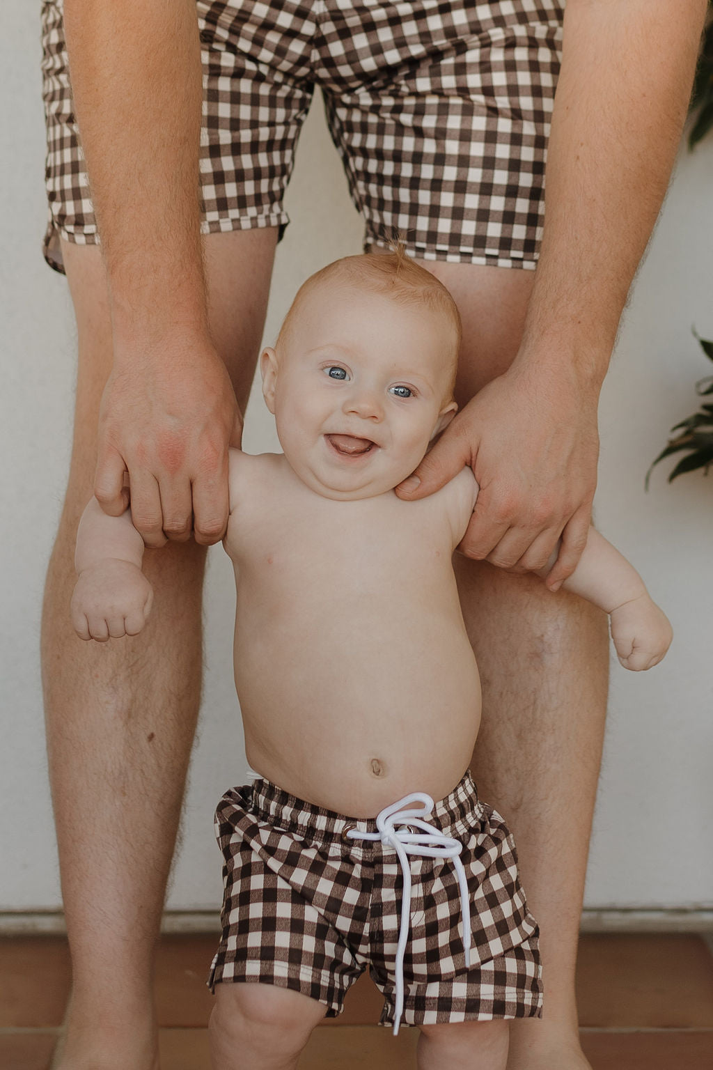 A smiling baby from the forever french baby Chocolate Gingham collection wears brown and white checkered shorts, held by an adult wearing matching Men's Boardshort | Chocolate Gingham. The tender moment unfolds indoors, with the adult's hands gently supporting the baby from behind.