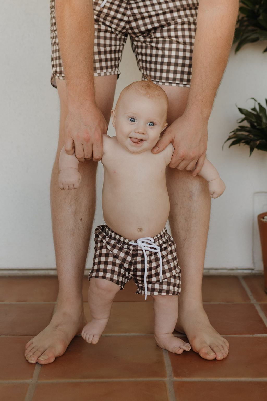 An adult's waist down holds a smiling baby on a tiled floor, both in matching Chocolate Gingham shorts by Forever French Baby, with lush plants enhancing the serene scene.