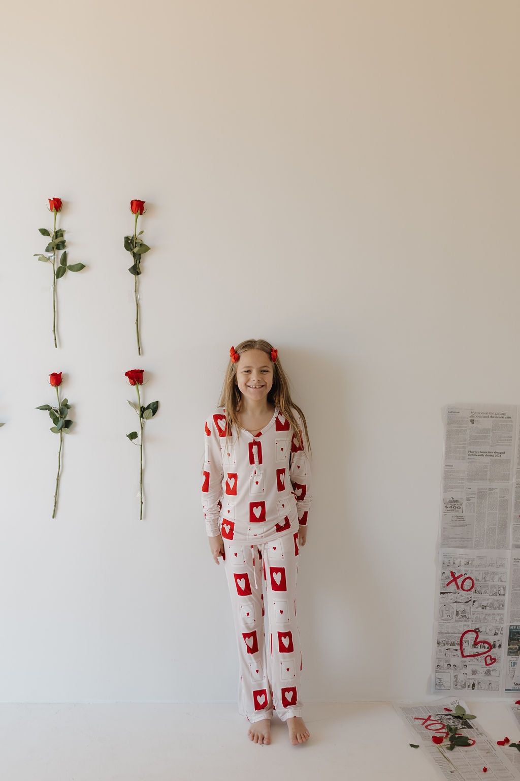 A smiling girl leans against a wall wearing forever french baby's Women's Bamboo Pajamas | Love Day, featuring red hearts. Roses are taped to the wall beside her, and a newspaper fragment lies on the floor.