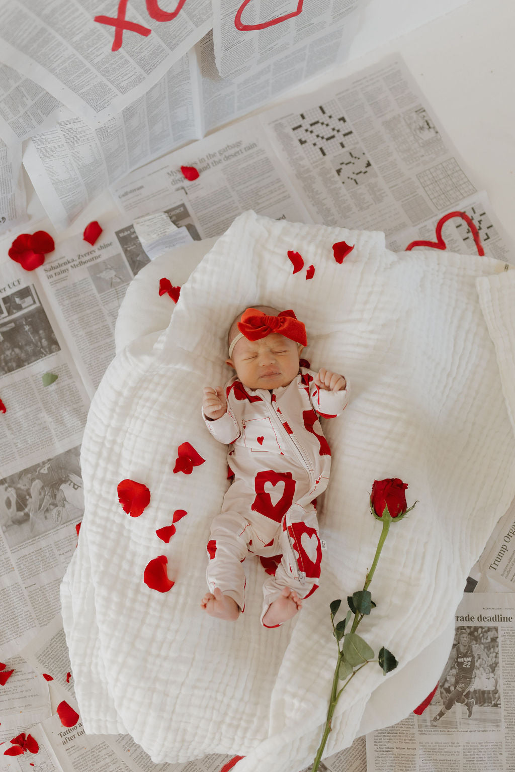 A baby in a "Love Day" bamboo zip pajama set by forever french baby sleeps on a breathable fabric blanket. They're adorned with a red headband, red rose petals, and a single red rose, all against a backdrop of newspaper sheets.