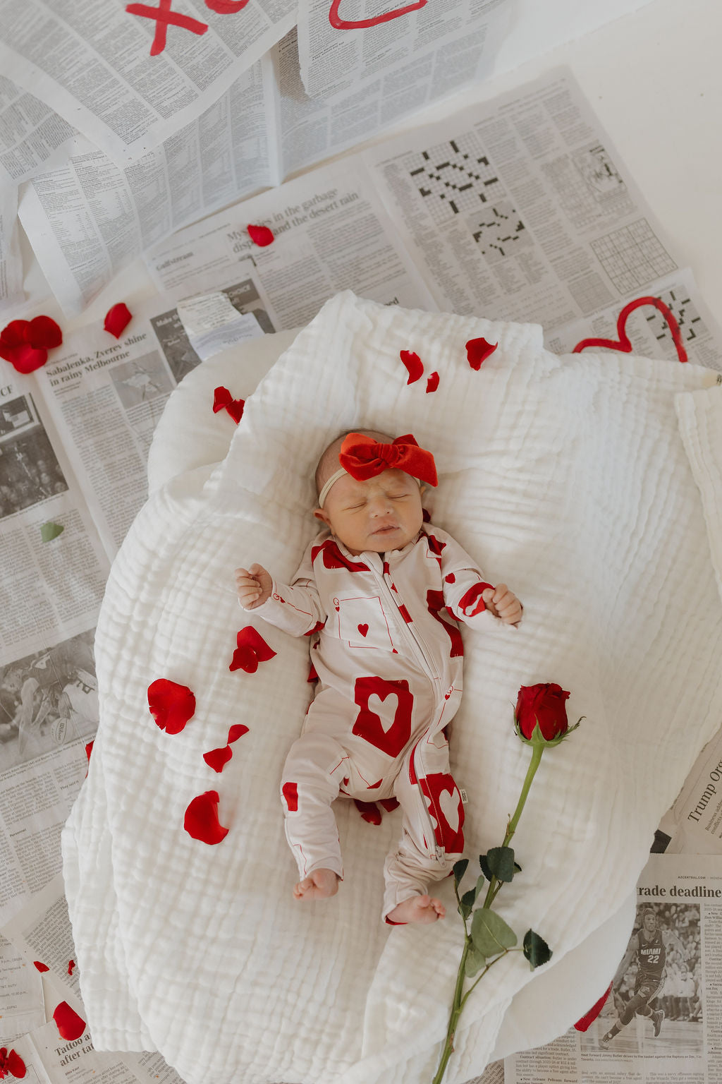 A baby peacefully sleeps on a white blanket covered with red rose petals, wearing "Bamboo Zip Pajamas | Love Day" by forever french baby. A matching bow headband and a single red rose beside the infant complete this serene scene set on newspapers.