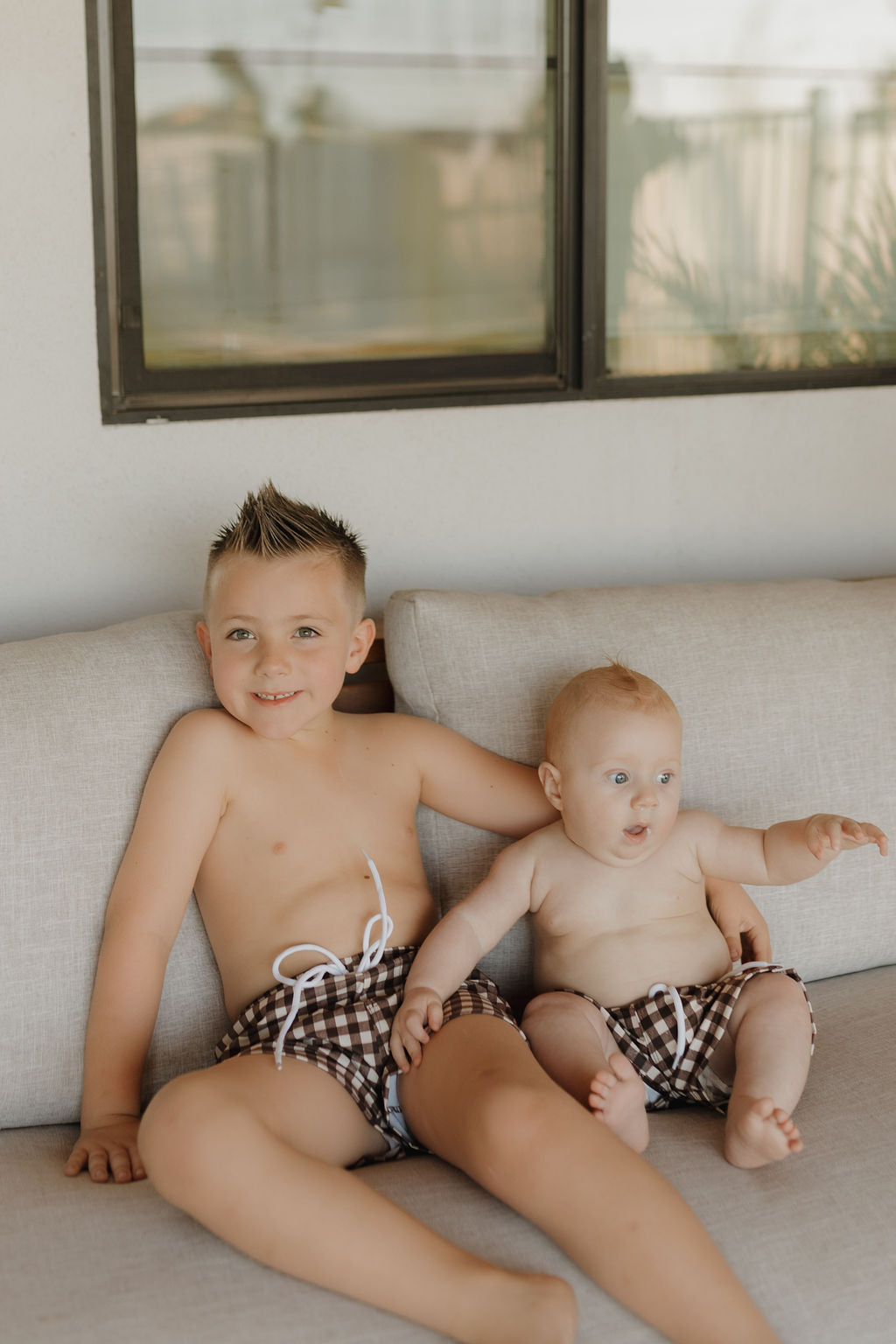 Two young children sit on a gray sofa in matching forever french baby Chocolate Gingham Child Boardshorts. The older child smiles, arm around the younger, who gazes off to the side. A window reveals plants outside, capturing the essence of Forever French Baby charm.