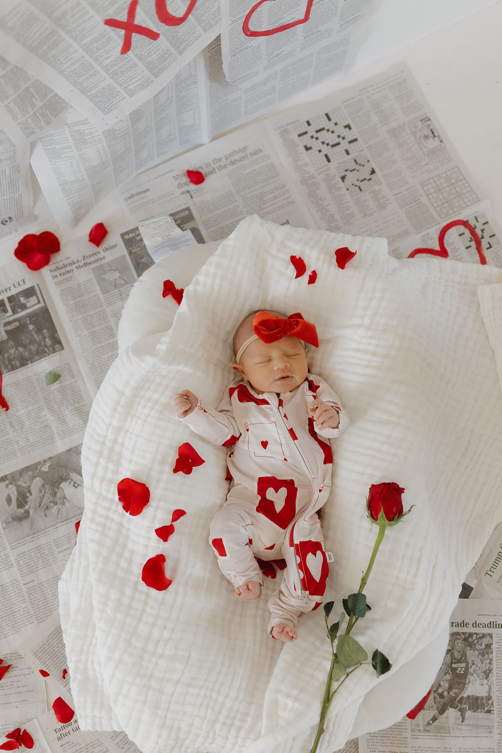 A baby in Love Day Bamboo Zip Pajamas by forever french baby and a large red bow headband lies on a white blanket with scattered red rose petals and newspapers. A single red rose rests beside the baby.