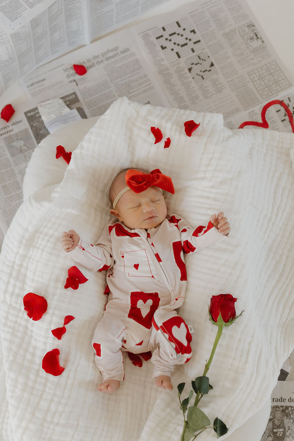 A baby in forever french baby's Bamboo Zip Pajamas | Love Day, featuring hypo-allergenic fabric with red hearts and a matching red headband, sleeps on a white blanket. Red rose petals are scattered around with a single red rose beside the baby, on newspapers underneath.