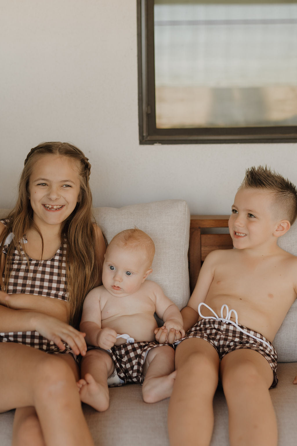 Three kids sit on a couch in swimsuits from the Chocolate Gingham Collection. The girl on the left, wearing a Forever French Baby piece with long hair and a brown checkered swimsuit, is joined by the baby and boy in matching trunks, all smiling against a sunlit window backdrop.