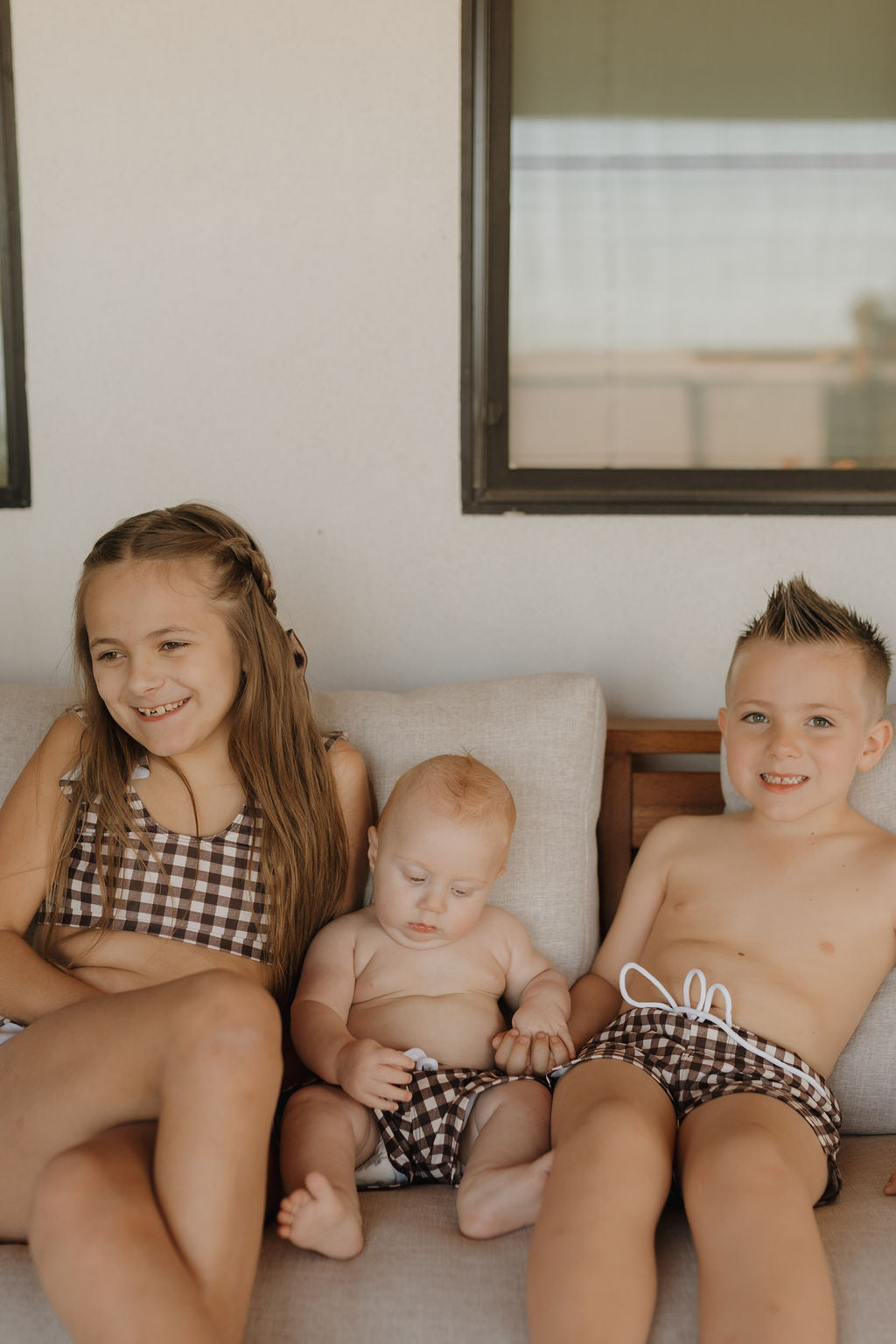 Three children in matching brown checkered swimwear from forever french baby sit on a couch. The older girl, in a Girls Two Piece Swimsuit | Chocolate Gingham, and boy smile, while the baby looks downward. A light wall and window backdrop emphasize their forever french baby charm.