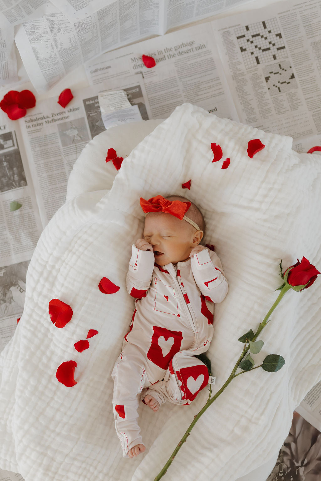 A baby sleeps on a white, textured blanket spread over newspapers, wearing forever french baby's Bamboo Zip Pajamas in the Love Day pattern and a red bow headband. Red rose petals and a single red rose are scattered around, enhancing the cozy and romantic vibe.