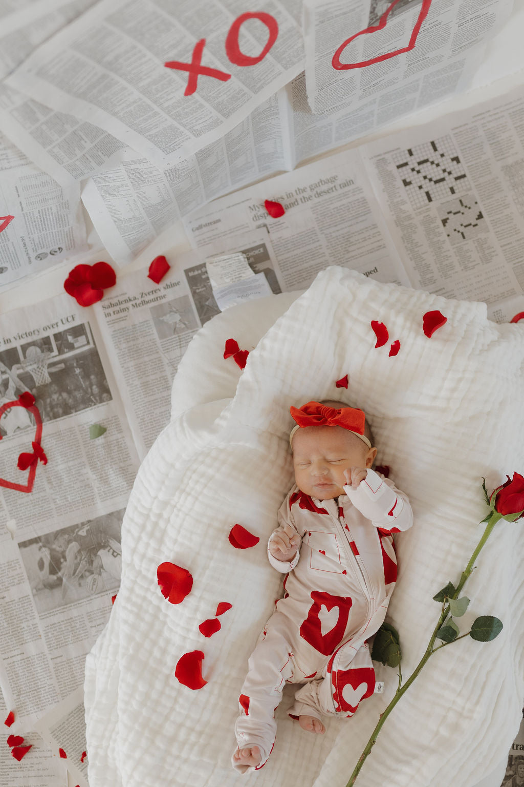 A baby rests on a white blanket, surrounded by newspapers and red rose petals. They're snug in forever french baby's Bamboo Zip Pajamas | Love Day, decorated with heart designs and paired with a red headband. The setting is warm with red roses and heart motifs.