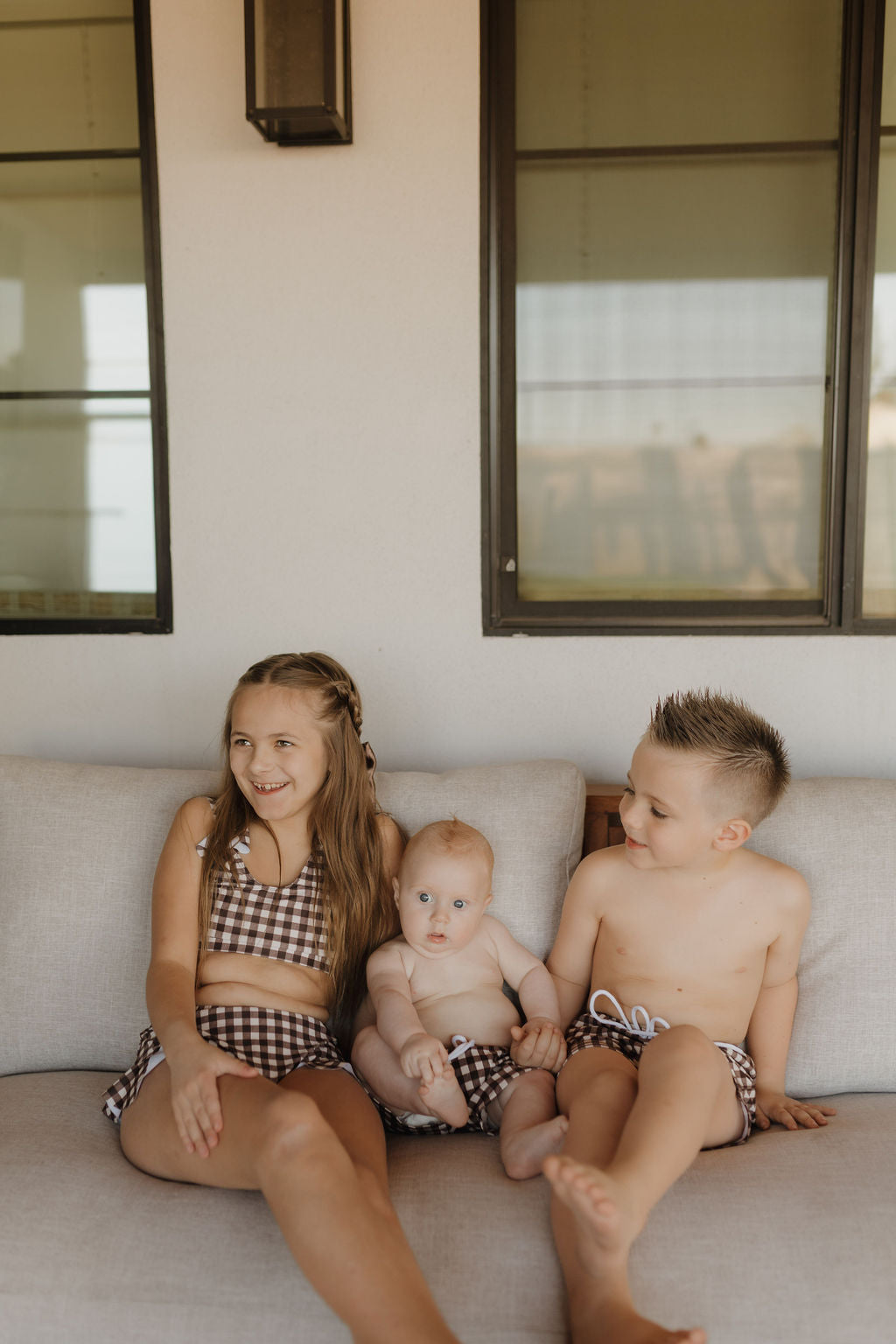 Three children in matching Chocolate Gingham swimwear from forever french baby sit on a light-colored sofa. The older siblings, seated at the sides, smile and look away while the baby in the center looks forward. Large windows are behind them.