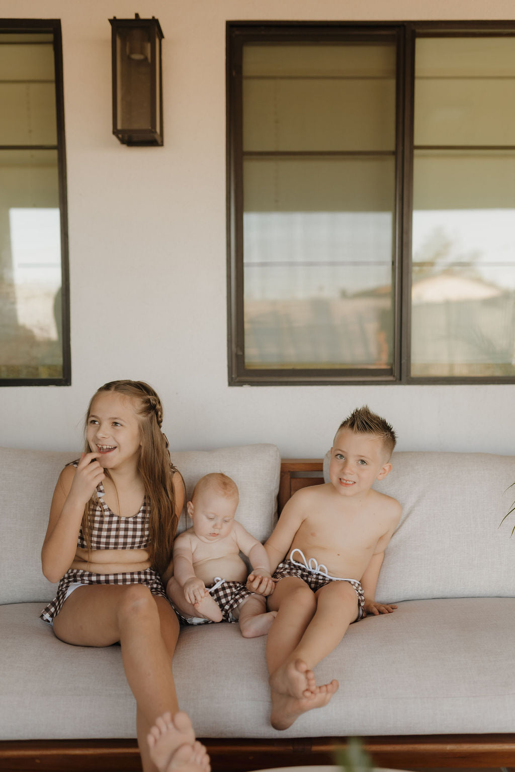 Three children sit on a couch in matching outfits from forever french baby’s Chocolate Gingham collection. The girl on the left smiles, the baby in the middle relaxes, and the boy on the right grins at the camera, with large windows creating a cozy indoor backdrop.