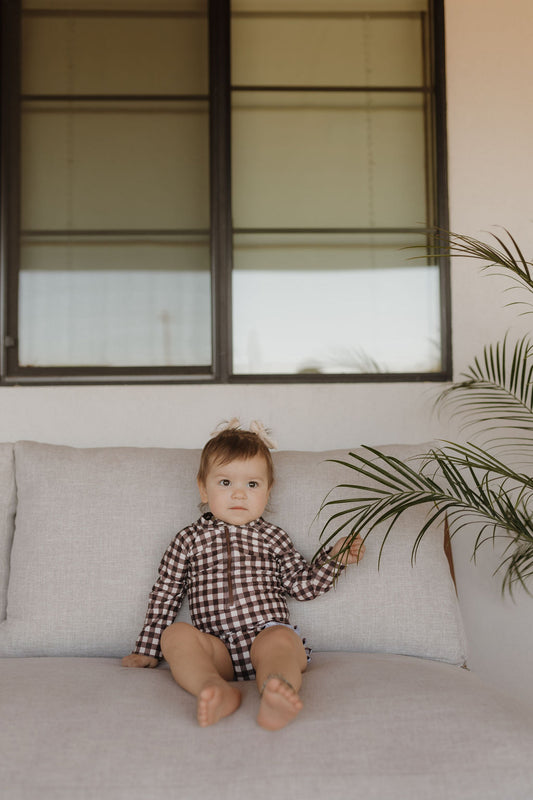 A small child, dressed in the Girls Long Sleeve Swimsuit from forever French baby's Chocolate Gingham Collection, sits on a light sofa. Holding a leaf, the scene is framed by a large window with dark edges.