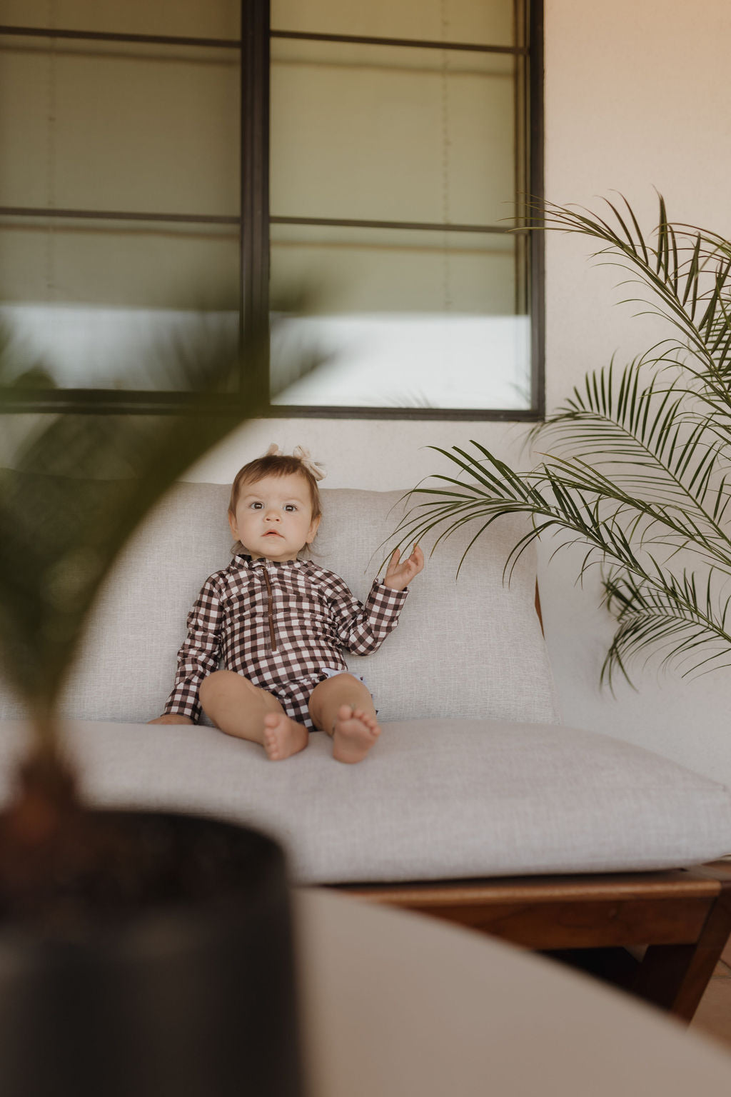 A toddler in a Girls Long Sleeve Swimsuit, Chocolate Gingham from forever French baby, sits on a light-colored sofa. They glance to the side with one hand raised, surrounded by lush green plants near a window.