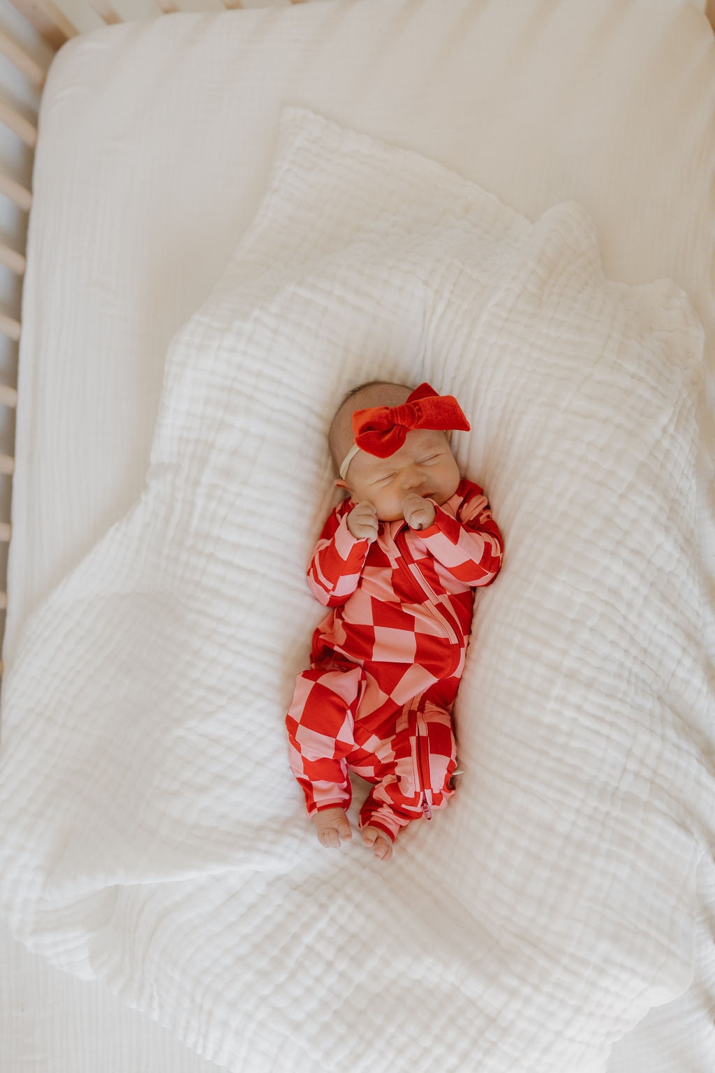 A baby rests on a white blanket, wearing breathable, hypoallergenic Bamboo Zip Pajamas | XOXO in red and white checkers by forever french baby, complete with a matching red headband. The crib mattress is layered with a soft, textured white sheet.