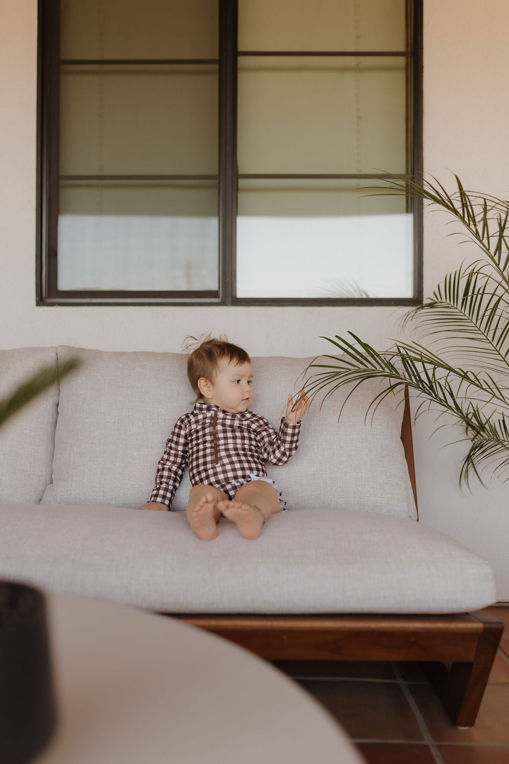 A child wearing a Girls Long Sleeve Swimsuit from the Chocolate Gingham Collection by forever French baby sits barefoot on a light sofa, gently touching a leafy plant beside them, with a window featuring dark frames in the background.