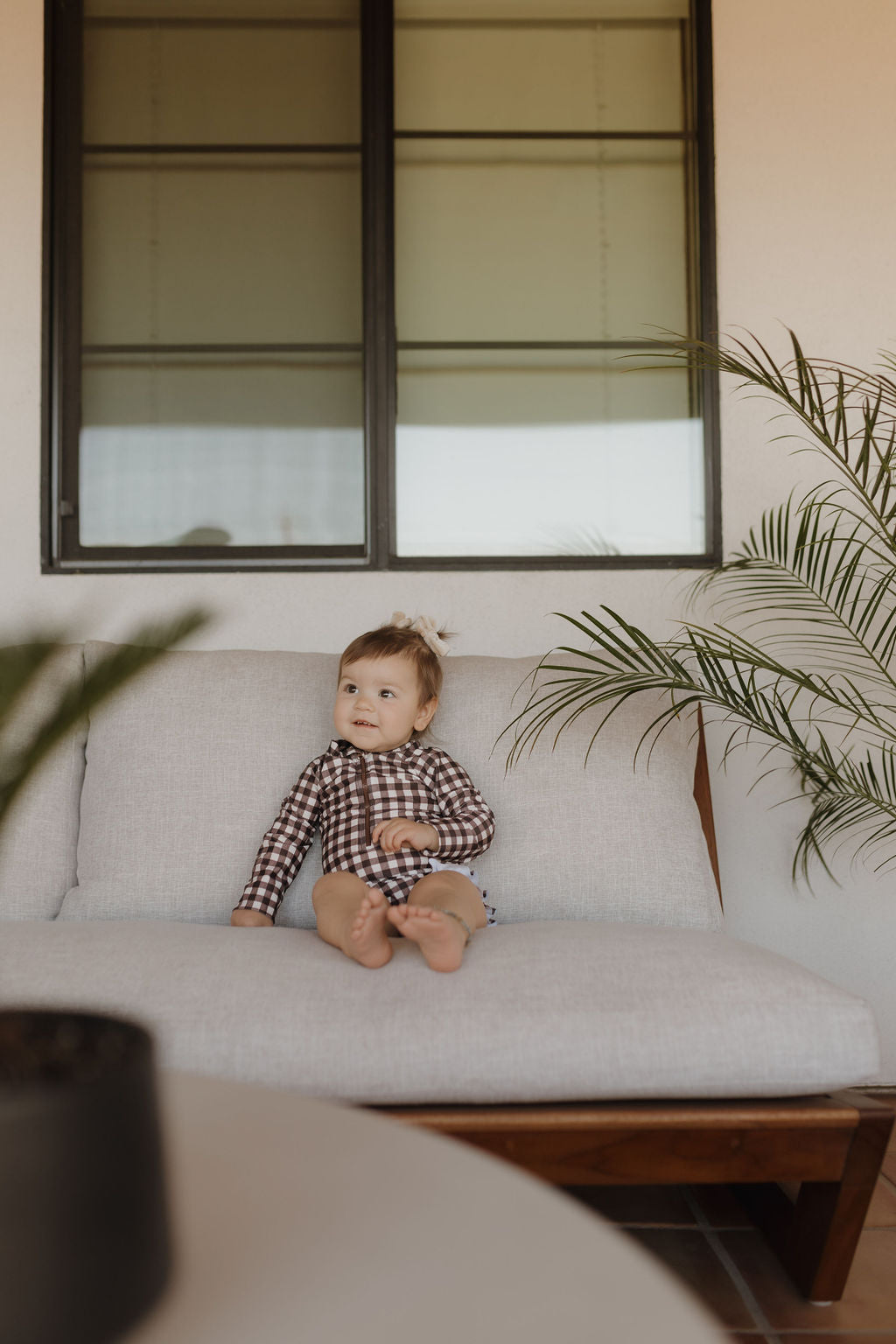 A baby, dressed in a Girls Long Sleeve Swimsuit from forever French baby's Chocolate Gingham collection, sits on a gray couch. Surrounded by plants and natural light streaming through a large window, this cozy scene embodies the timeless charm of Forever French Baby style indoors.