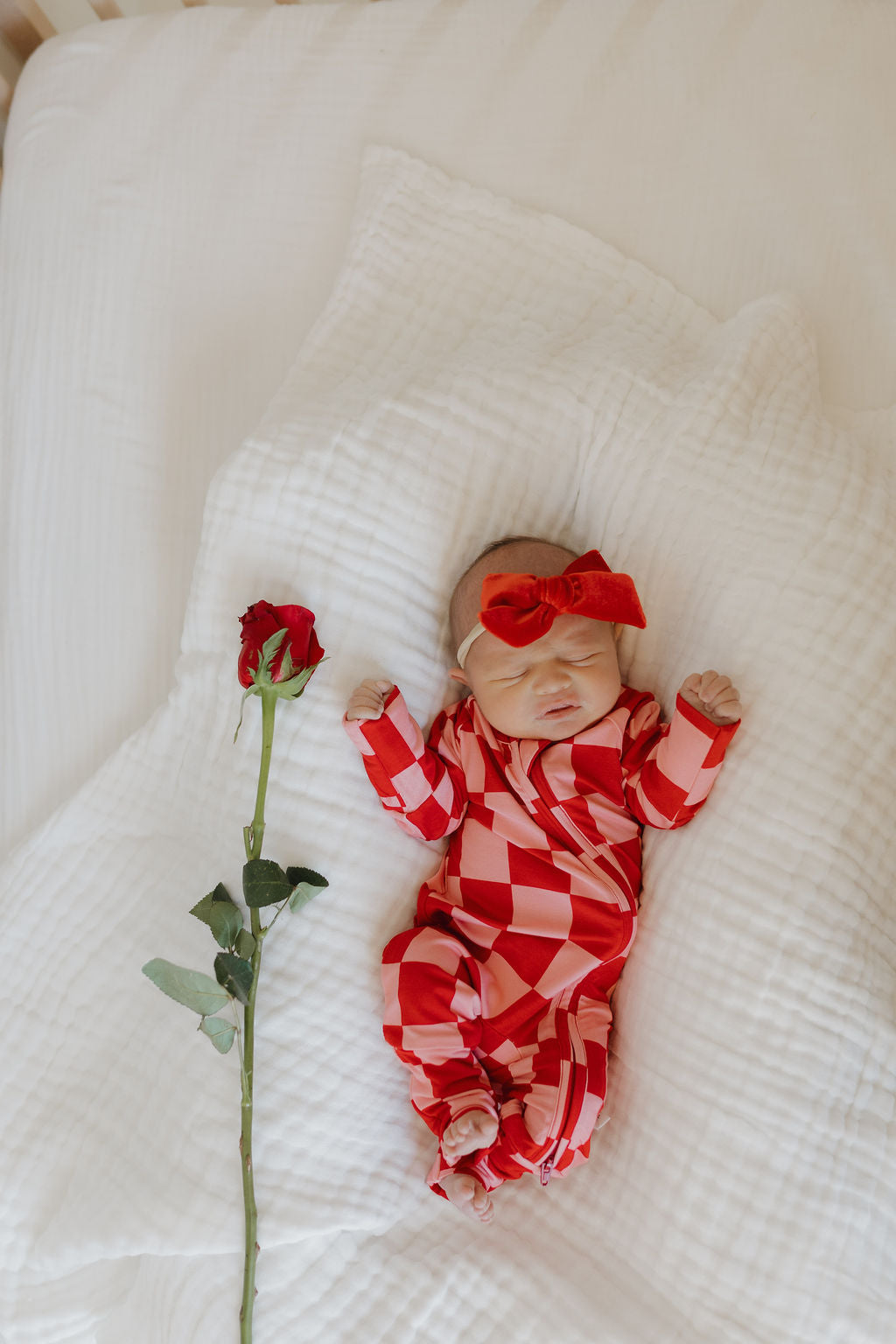 A newborn rests on a white blanket in a red and white checkered Bamboo Zip Pajamas by forever french baby, paired with a matching red headband. A single red rose lies beside the charming little one.