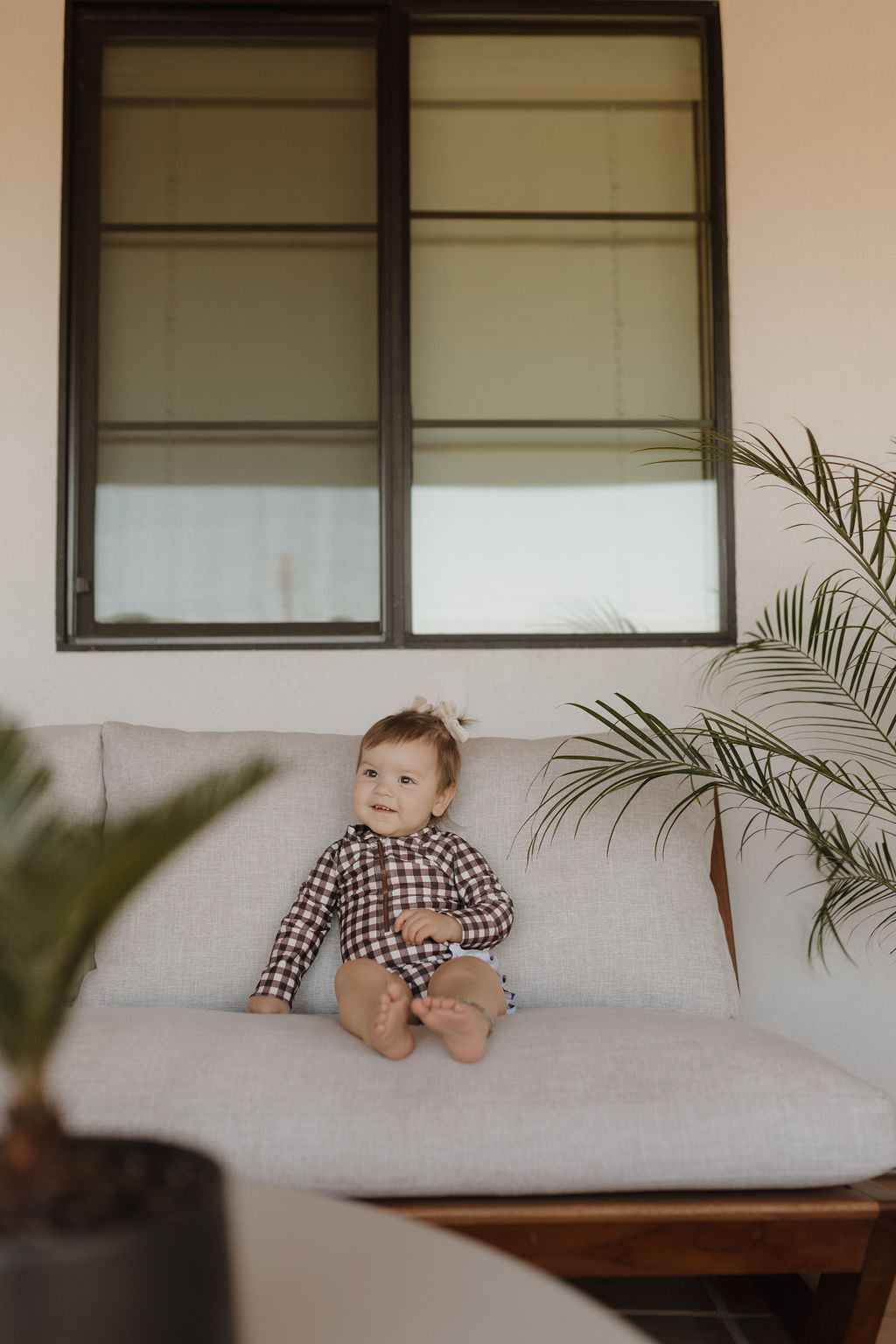 A baby in a Girls Long Sleeve Swimsuit from Forever French Baby's Chocolate Gingham Collection sits on a gray padded bench by a large window, with nearby potted plants enhancing the modern setting and timeless elegance of the brand.