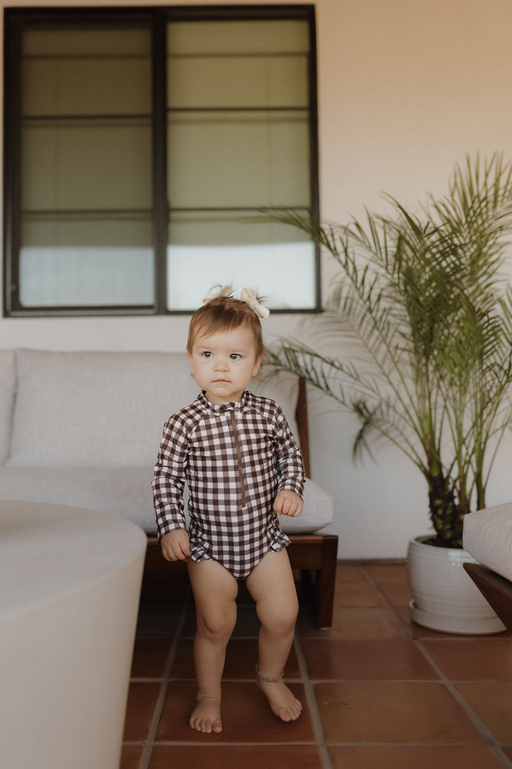 A toddler with short brown hair stands on terracotta tiles in a checkered outfit from the Forever French Baby Chocolate Gingham collection. A cozy sofa and potted plant sit against a wall with a large window, embodying the brand's style.