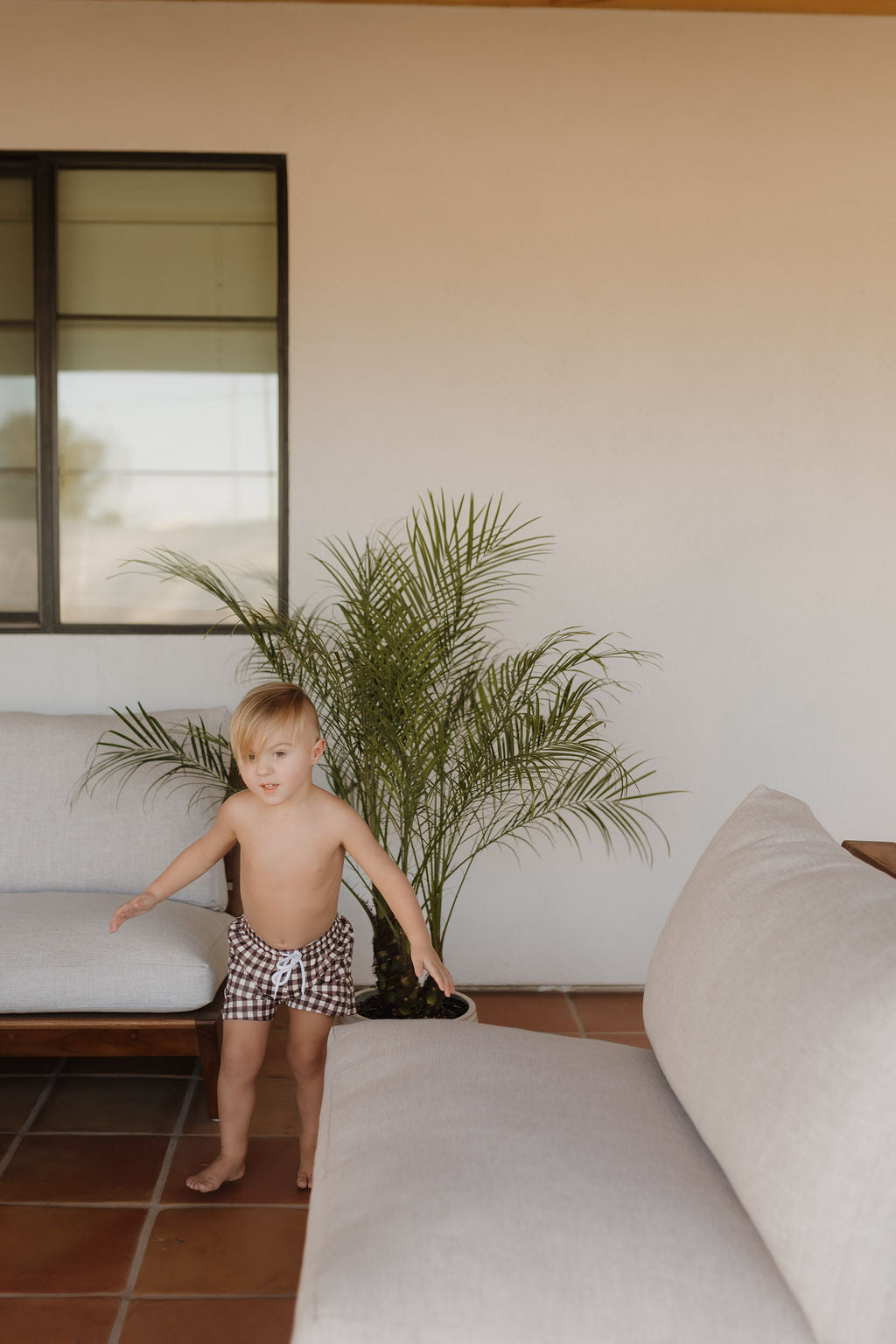 A young blond-haired child stands barefoot on a tiled patio, wearing plaid shorts from the forever french baby Chocolate Gingham Child Boardshort collection, beside a small potted plant, with a large window, white wall, and light outdoor seating in the background.