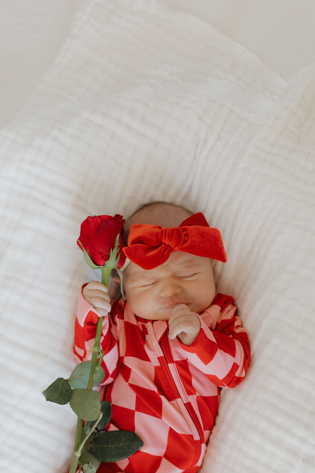 A baby is peacefully lying on a white quilt, dressed in breathable "Bamboo Zip Pajamas | XOXO" by forever french baby, with a matching red bow headband and holding a single red rose.