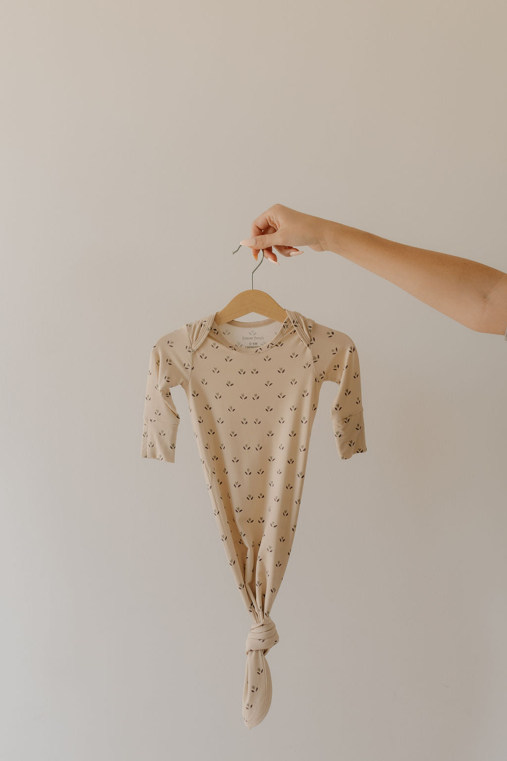 A hand holds a bamboo "Flora" knotted gown from forever french baby, designed for newborns. It's beige with subtle black motifs and hangs on a wooden hanger against a light-colored wall. Hypoallergenic and essential, it's crafted from eco-friendly materials.