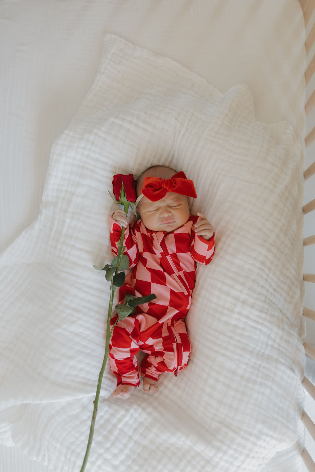 A newborn peacefully sleeps on a white blanket in a crib, wearing breathable Bamboo Zip Pajamas XOXO by forever french baby, with matching headband and tiny fists raised near its head, holding a long-stemmed red rose.