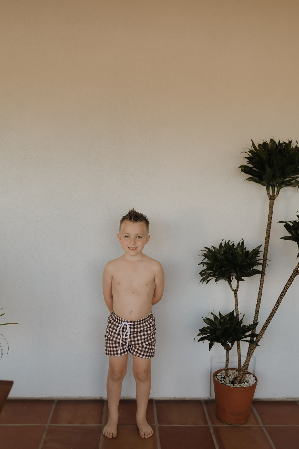 A young boy stands barefoot on a tiled floor wearing Child Boardshorts in Chocolate Gingham by forever french baby. He smiles slightly against a plain wall, with two potted plants beside him—one large with broad leaves and one smaller—in a bright, minimalistic setting.