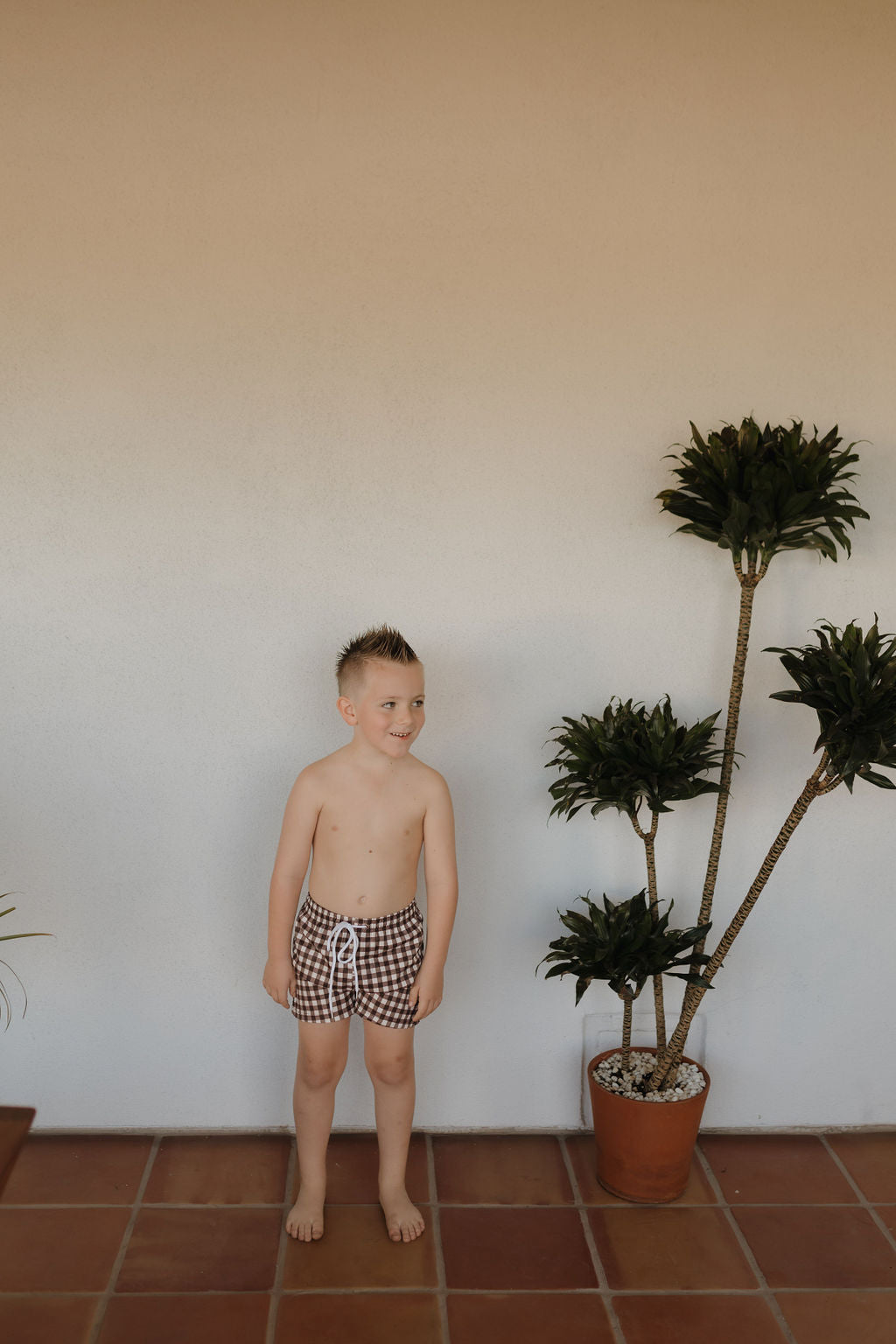 A young boy smiles barefoot indoors on a tiled floor, wearing Forever French Baby's Child Boardshort in Chocolate Gingham. Standing next to a leafy potted plant against a plain, light wall, he embodies the brand's carefree spirit.