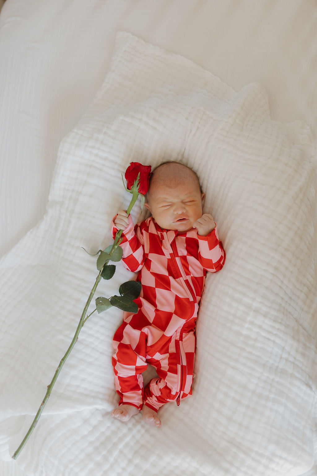 A newborn rests serenely on a white blanket, clad in breathable Bamboo Zip Pajamas by forever french baby, featuring a red and pink checkered design. The baby holds a single red rose, nestled beside them on the soft fabric.