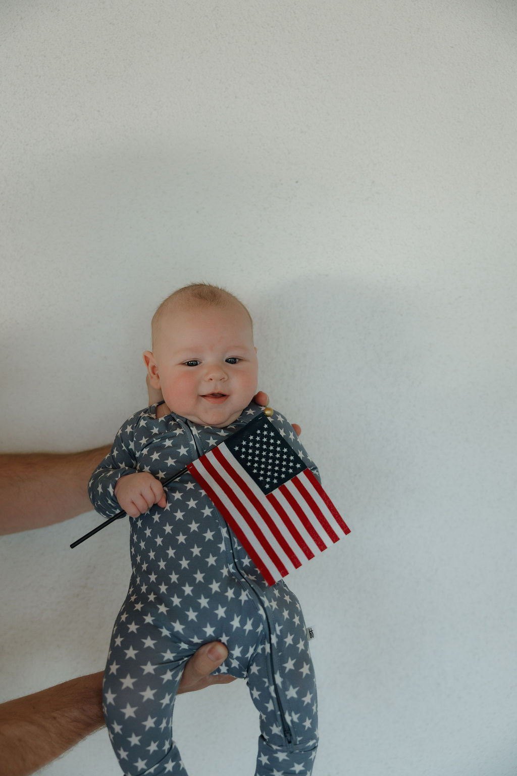 A smiling baby in forever french baby Bamboo Zip Pajamas | Star Spangled, featuring a star pattern, is held by an adult’s hand and holds a small American flag against a plain white background.
