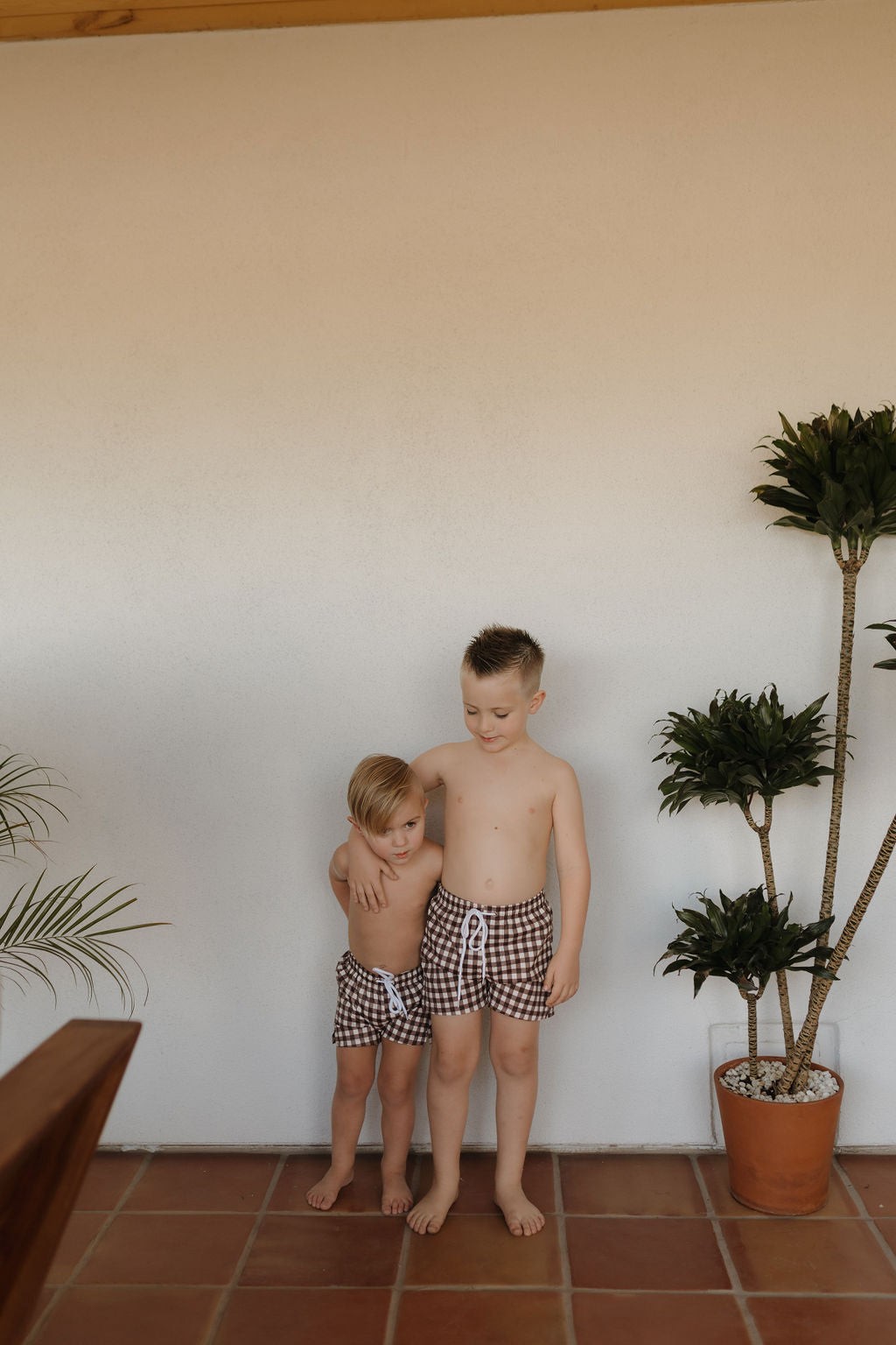 Two young children stand on a tiled floor wearing the Child Boardshort in Chocolate Gingham from forever french baby. The older child has an arm around the younger, with potted plants on either side, creating a timeless scene.