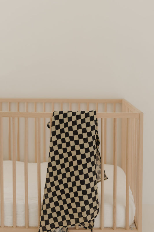 A light wooden crib with a white mattress sits against a plain beige wall. A forever french baby Muslin Swaddle | Black Checkerboard drapes over the crib’s side.