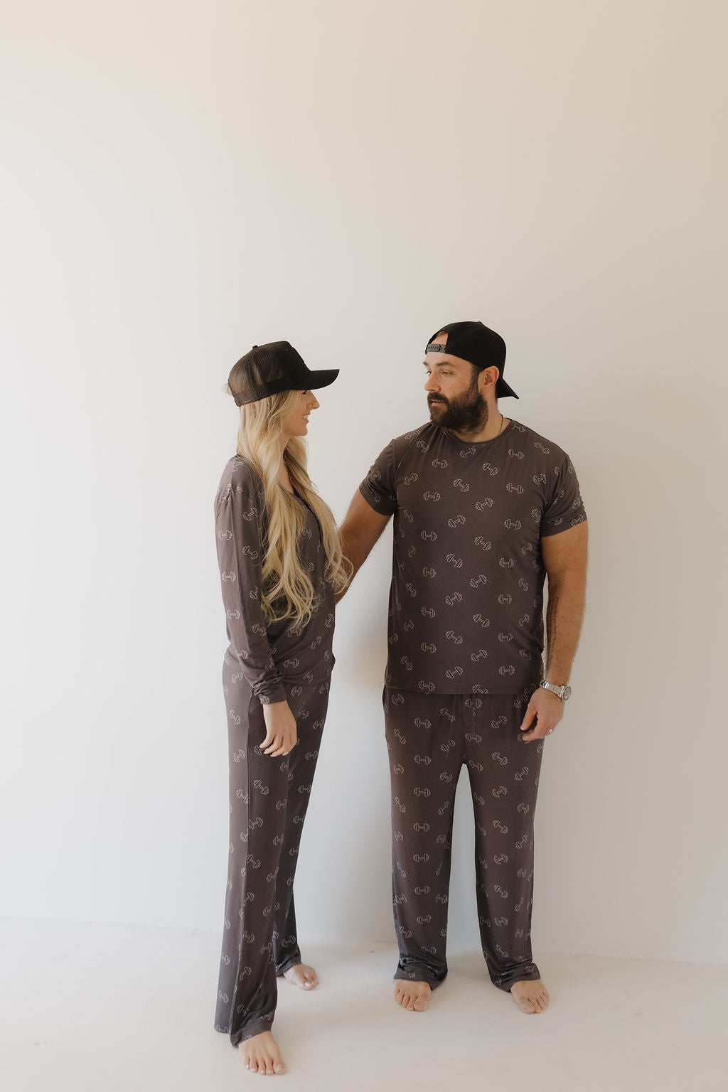 A man and woman wear matching forever french baby outfits, both in the Bamboo Men's Short Sleeve Pajama | Charcoal Arm Day. They're barefoot against a plain background, donning black caps. He gazes at her as she looks back.