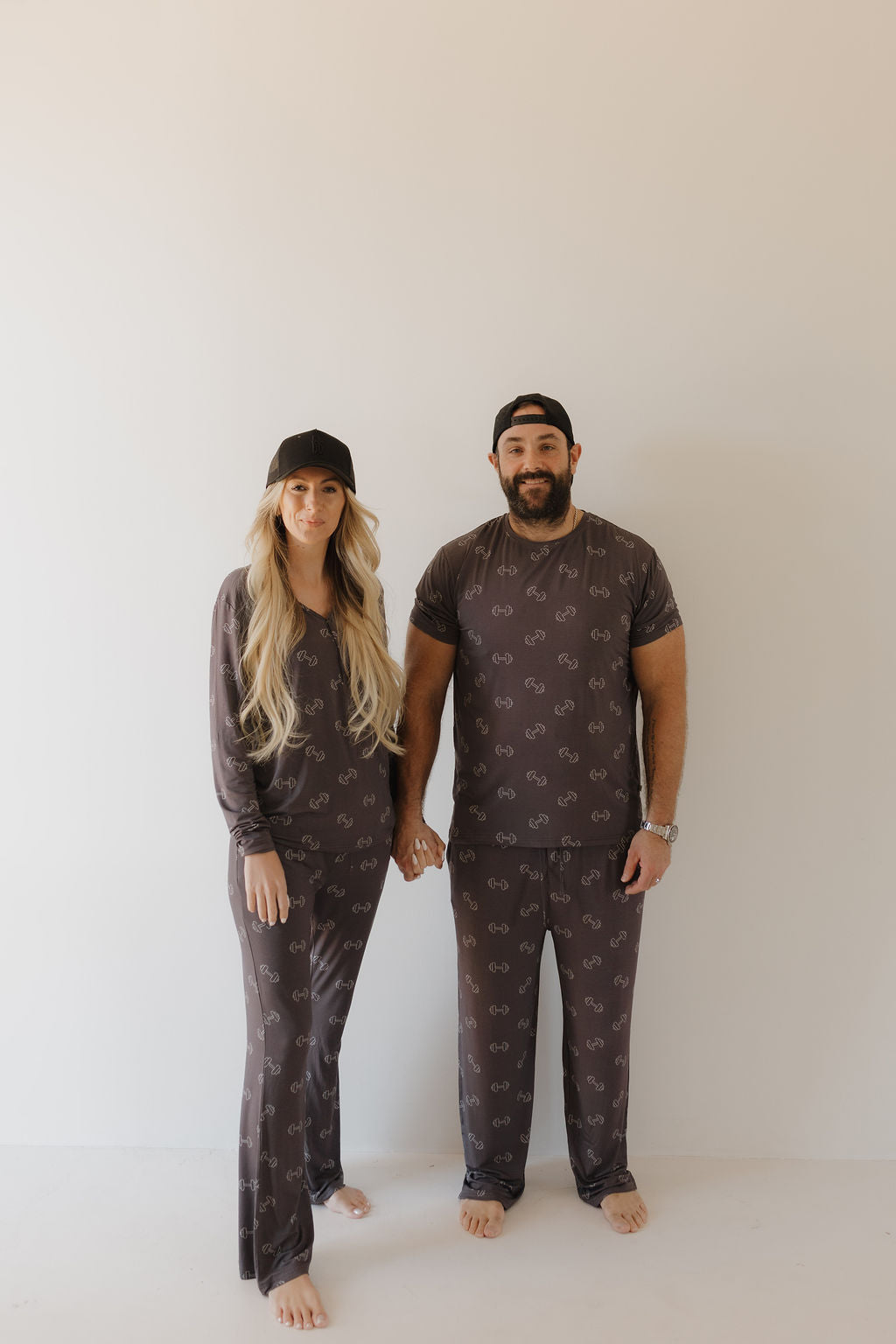 A couple stands barefoot on a plain background in matching forever french baby sleepwear, holding hands. The bearded man and blonde woman both wear black baseball caps and Bamboo Men's Short Sleeve Pajamas in Charcoal Arm Day, ensuring comfort with their hypoallergenic fabric.