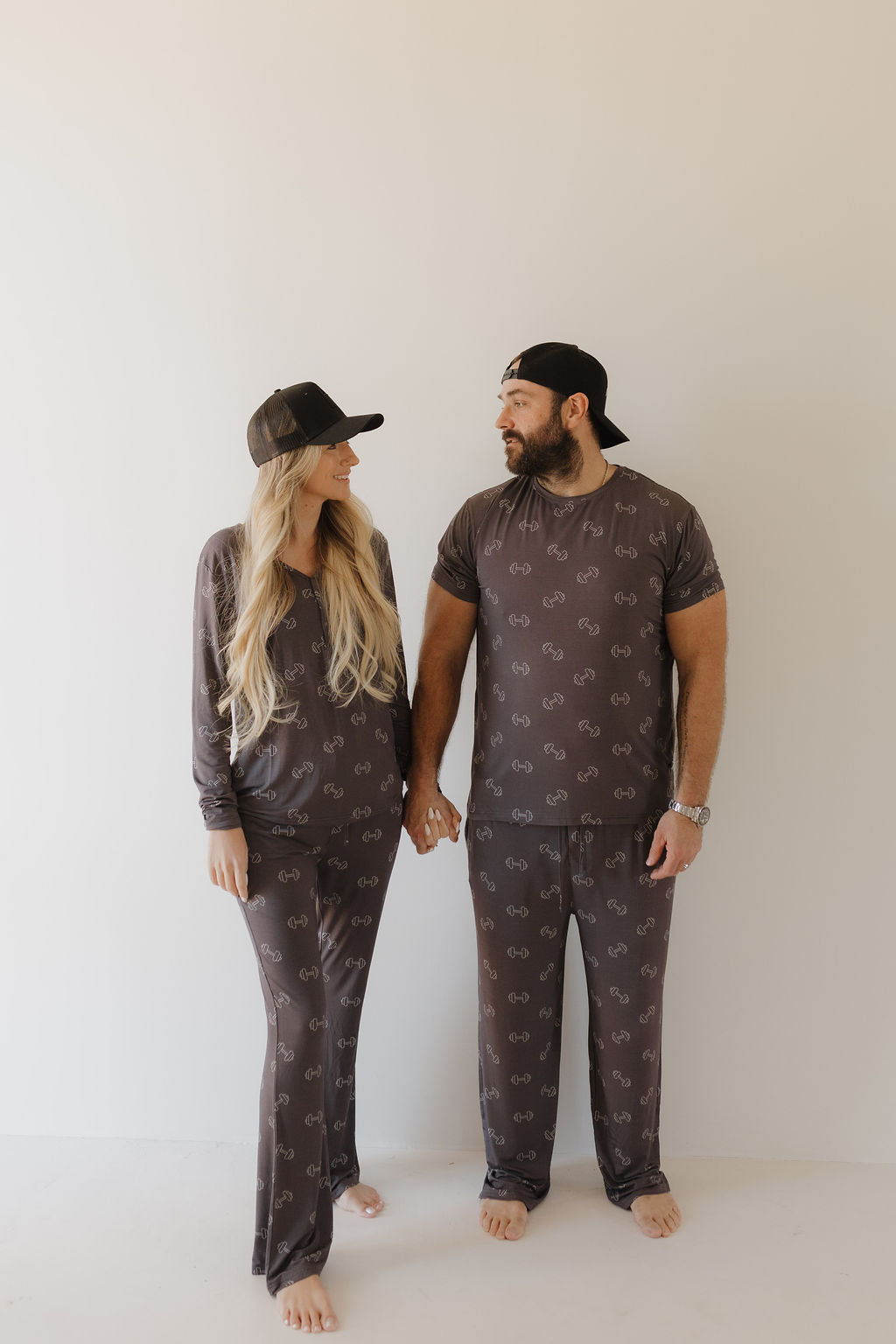 A couple in matching Women's Bamboo Pajamas | Charcoal Arm Day by forever french baby stands barefoot against a white background. They both wear black baseball caps, holding hands and gazing at each other.