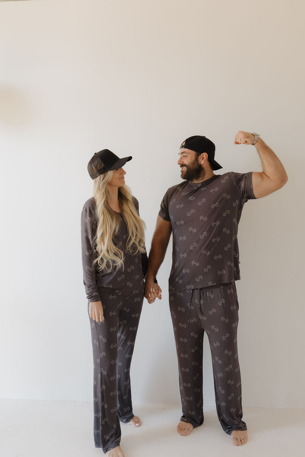 A couple stands holding hands against a plain backdrop, both in matching gray pajamas. The man, wearing a backward cap, flexes playfully while looking at the woman in forever french baby's Women's Bamboo Pajamas - Charcoal Arm Day and a forward cap, both smiling.