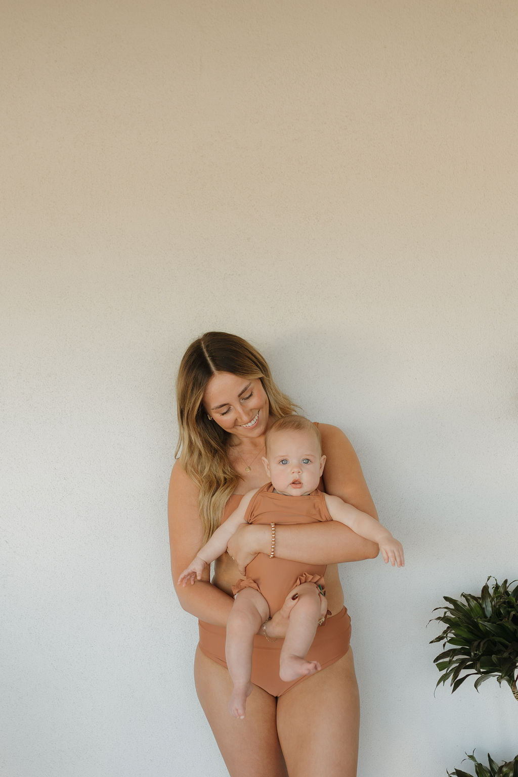 A woman smiles while holding a baby, both in coordinating brown outfits from the forever french baby Terra collection. They stand against a plain wall with a leafy plant partially visible on the right.