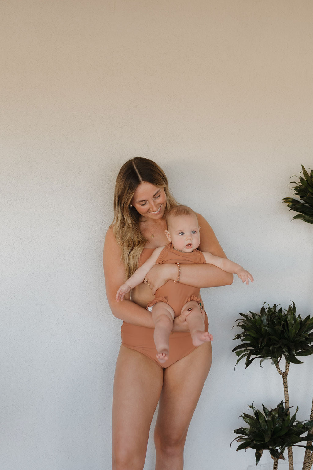 A woman wearing a Women's Two Piece Swimsuit from the Terra collection by forever french baby smiles while holding a baby in a matching outfit, standing against a plain wall with small potted plants nearby.