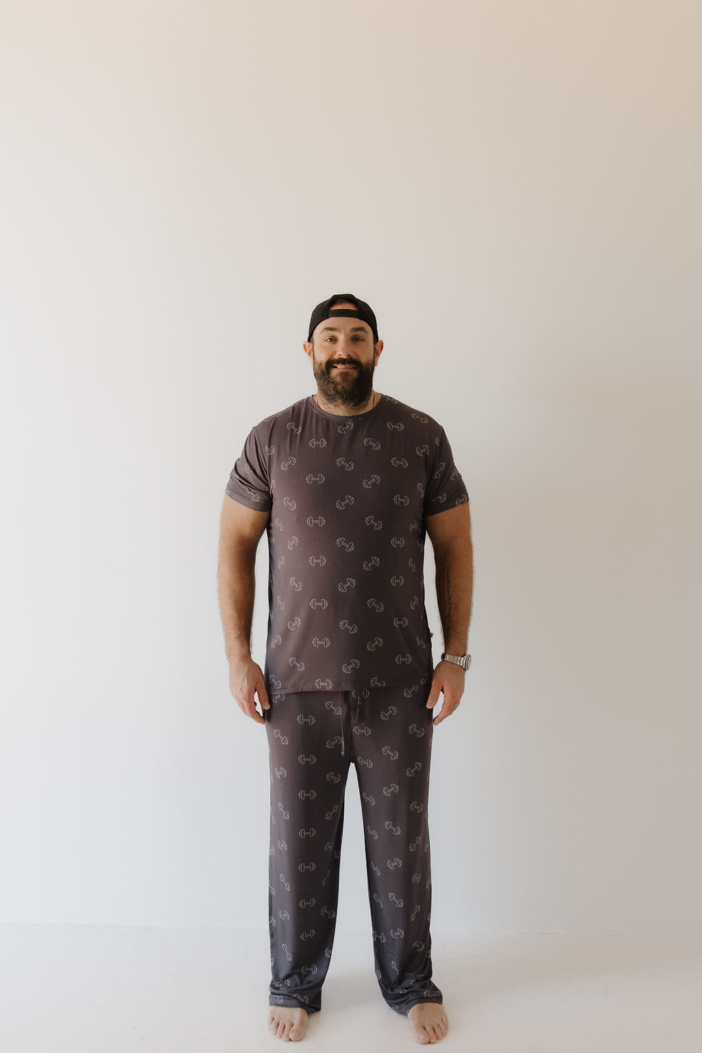 A bearded man, barefoot against a white backdrop, wears "Bamboo Men's Short Sleeve Pajama | Charcoal Arm Day" by forever french baby. Dressed in these hypo-allergenic pajamas with circular patterns and a black cap, he embodies both comfort and style.