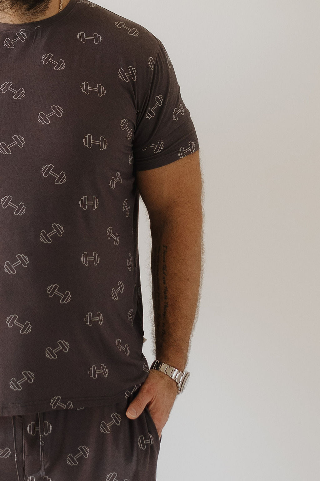 A person wearing the forever french baby Bamboo Men's Short Sleeve Pajama in Charcoal Arm Day, with a dumbbell design, is shown from shoulders to waist. The breathable fabric ensures comfort. A tattoo adorns their forearm, and they wear a watch while resting a hand casually in their pocket.