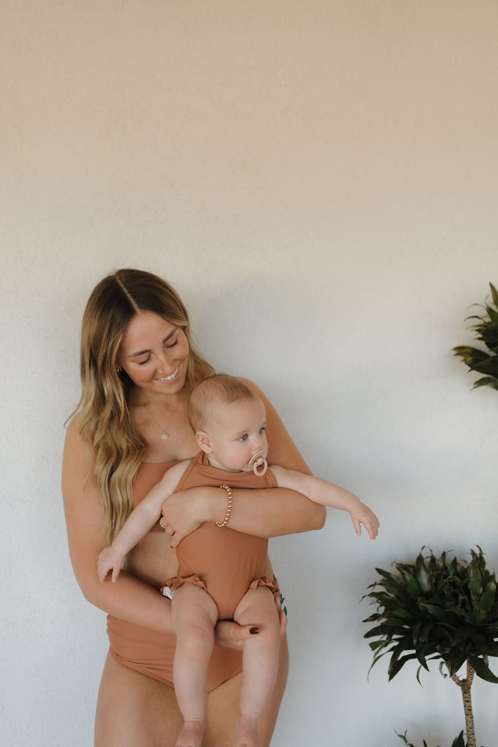 A woman with long hair holds a baby dressed in a matching brown outfit from the Terra collection by Forever French Baby. They stand before a light-colored wall framed by green plants, both gazing calmly to the left.